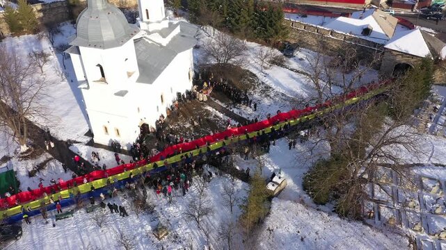 Static aerial close-up of a large crowd holding a massive, extended Romanian flag, tricolor on the snow-covered grounds of Raducanu Monastery in Targu Ocna during a winter national celebration.