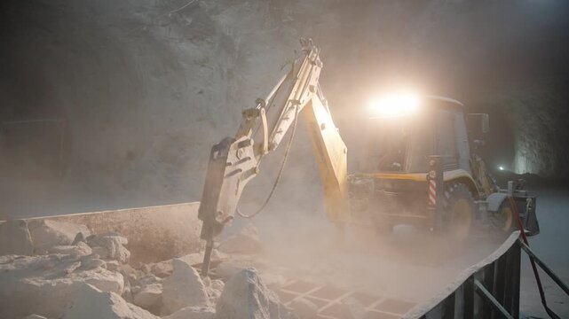 Static, close-up shot of a backhoe loader using a hydraulic breaker to break white rock inside a massive, dimly lit, dust-filled underground mine cavern.