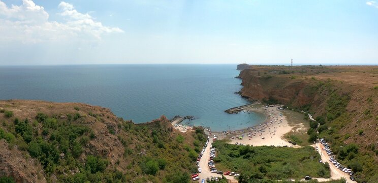 Aerial drone image of Bolata Beach, Bulgaria, showing the curved bay with red vertical cliffs, pier, summer sea, sunbathing tourists, and Cape Kaliakra visible in the distance