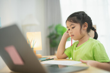 Obraz premium Young girl focusing on her homework with a laptop, colorful stationery, and a cozy study space, engaged in online learning at home