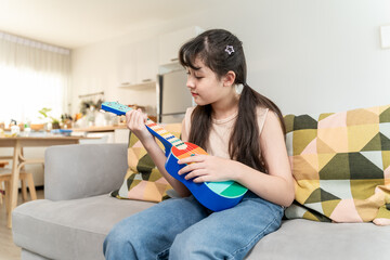 Asian young girl sitting on sofa and playing an ukulele in house. 
