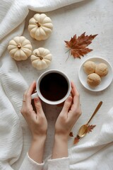 Cozy autumn flat lay with female hands holding a warm cup of coffee surrounded by mini white pumpkins, cookies, a golden spoon, and a dry maple leaf on soft fabric background in warm seasonal atmosphe