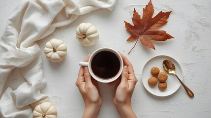 Cozy autumn flat lay with female hands holding a warm cup of coffee surrounded by mini white pumpkins, cookies, a golden spoon, and a dry maple leaf on soft fabric background in warm seasonal atmosphe