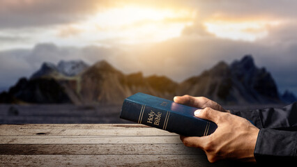 Prayer holding the blue book of the holy bible on a wooden table with a landscape view and a dark...