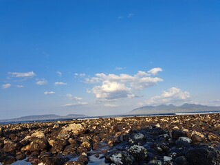 Rocky shoreline with tide pools under a bright blue sky with scattered clouds, distant mountains visible on the horizon.