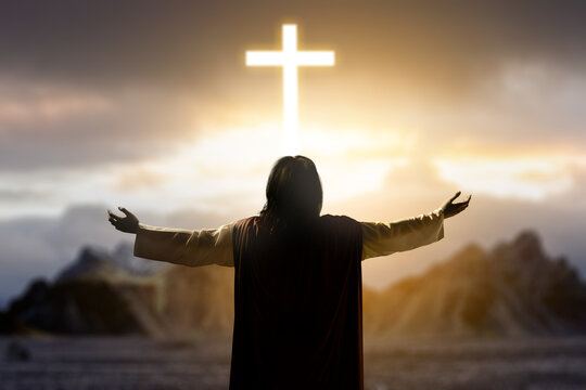 Rear view of Jesus Christ raising his hand and praying with the light of the Christian cross on the hill with dark dramatic sky background. Concept of Christianity, religious, faith, Jesus, or belief