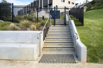 Concrete staircase with stainless steel handrails leading up to a black metal security fence and gate outside a modern apartment complex in Australia. Secured contemporary urban residential complex.