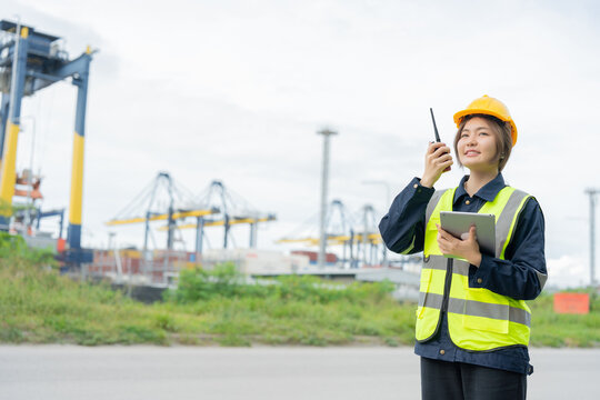 A female engineer stands confidently near towering cranes at the port, reviewing technical data on her tablet and coordinating with the control center to maintain smooth logistics operations.