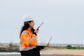 A woman wearing an orange jacket and a white helmet is holding a walkie talkie. Female engineer or supervisor in safety helmet and PPE at an industrial construction site or energy plant. © VStudio
