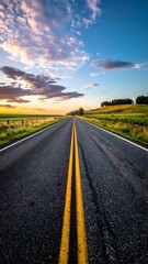Naklejka premium A scenic, straight road with double yellow lines stretches toward the horizon under a vibrant sunset sky with scattered clouds and rolling green hills