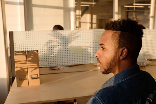 Profile portrait of young African American man in denim shirt sitting at office table and watching sunset thoughtfully