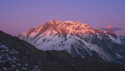 Majestic snow-capped mountain range stands beneath a vibrant, colorful sky during a scenic sunrise, with the mountain peaks illuminated by the warm, golden light of dawn reflecting off the icy...