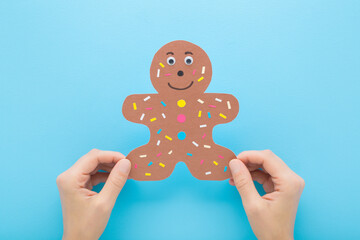 Woman hands holding brown gingerbread man shape from application paper on light blue table background. Pastel color.  Making decoration elements. Closeup. Point of view shot. Flat lay. Top down view.