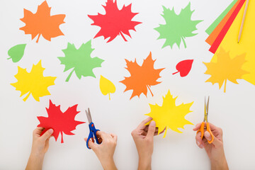 Young adult mother and little child hands cutting colorful maple leaves from application paper on light gray table background. Closeup. Making beautiful autumn decoration. Flat lay. Top down view.