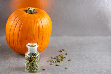 Glass jar filled with pumpkin seeds and whole pumpkin on gray surface, with scattered pumpkin seeds...