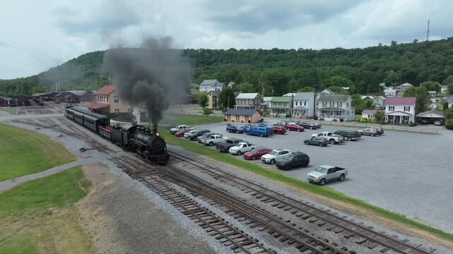 A narrow gauge steam train puffs clouds of smoke as it moves past a small town. The engine is surrounded by parked cars and charming houses under a bright sky.