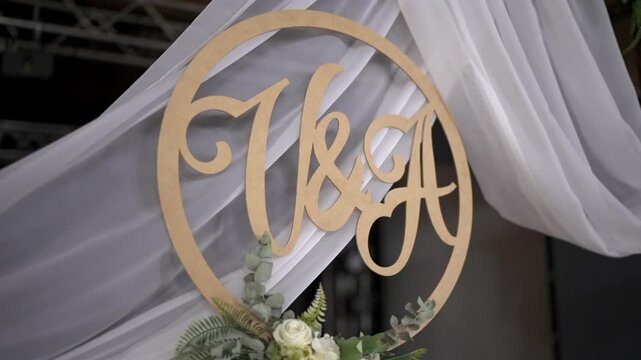 Decorative wooden monogram &ldquo;V and A&rdquo; framed in a circle, set against drapery and a floral base of white roses, eucalyptus, and ferns &mdash; ideal for weddings or anniversaries.