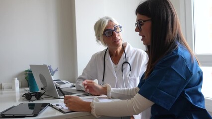 Experienced general practitioner training a younger female colleague, discussing a patient's medical history and treatment plan while reviewing documents and data on a laptop in the hospital office - Powered by Adobe