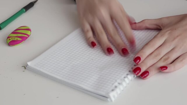 Woman using white rubber eraser to remove pencil marks from grid paper notebook, correction process with red manicured nails visible in detail.