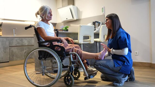 Female caregiver providing physical therapy and foot massage to an elderly woman in a wheelchair during a home visit, assisting with rehabilitation, mobility, and pain relief for the patient - Powered by Adobe