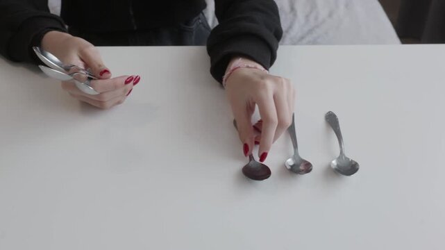 Woman with red nail polish placing silver tea spoons on desk surface in organized pattern, setting table or sorting cutlery activity.