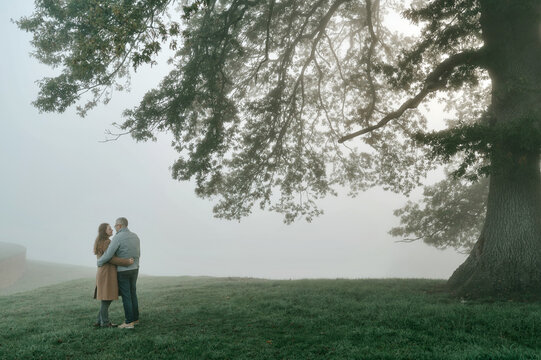Couple standing together under a large tree in foggy landscape.