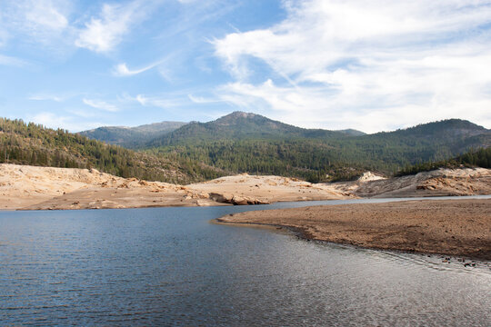 Receding water reveals sandy lakebed beneath forested hills