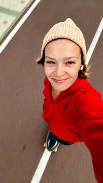 Woman in beanie and red hoodie smiling on skateboard, street selfie