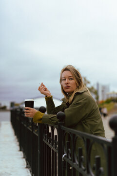 Woman leaning on black railing holding coffee and smoking a cigarette