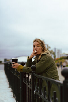 Woman leaning on black railing holding coffee and smoking a cigarette
