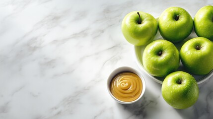 Green apples arranged in a bowl next to a small dish of caramel sauce on a marble surface.