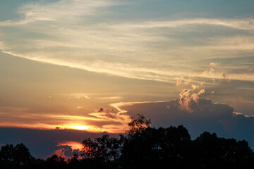 Fiery summer sunset paints the sky with vivid orange and gold hues, highlighted by soft clouds and dark tree silhouettes