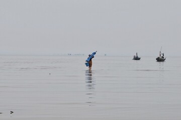 Naklejka premium Fisherman Wading in Shallow Sea with Small Boats in Misty Horizon Under Gray Sky