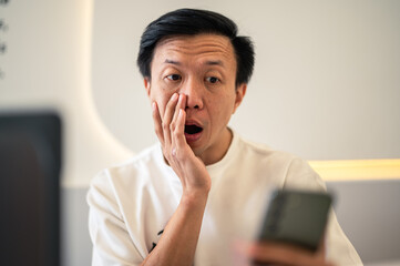 Asian man sitting at a café table, looking shocked while staring at his smartphone screen, reacting to surprising or unexpected news with an expressive face in a modern indoor workspace setting