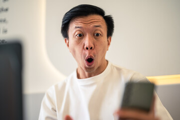 Asian man sitting at a café table, looking shocked while staring at his smartphone screen, reacting to surprising or unexpected news with an expressive face in a modern indoor workspace setting