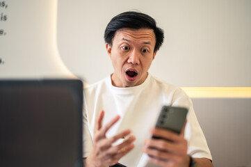 Asian man sitting at a café table, looking shocked while staring at his smartphone screen,...
