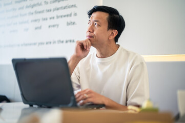 Asian man sitting at a modern café table, resting his chin on his hand and looking up thoughtfully while working on a laptop, wearing a white shirt in a bright, minimalist indoor setting.