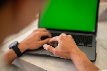 Fototapeta premium Close-up of an Asian man’s hands typing on a laptop with a green screen at a café table, showing modern workspace setup and technology focus in a professional environment