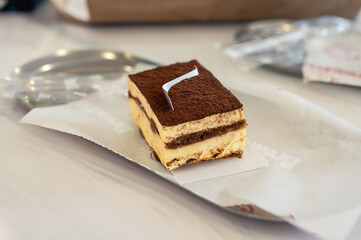 Close-up of a delicious slice of tiramisu cake with layers of cream and cocoa powder on top, served on white parchment paper over a marble table in a cozy café setting.