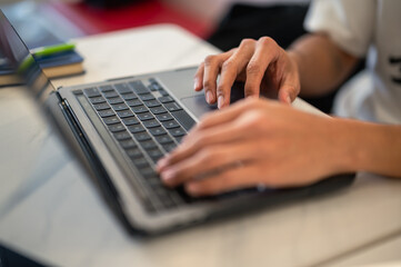 Close-up of an Asian person’s hands typing on a laptop keyboard at a café table, showing focus and productivity in a modern workspace environment with natural light and minimal design