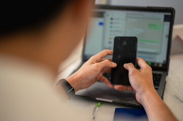 Asian man using a smartphone while sitting in front of a laptop with a messaging app on the screen, multitasking between phone and computer in a modern workspace with blurred background