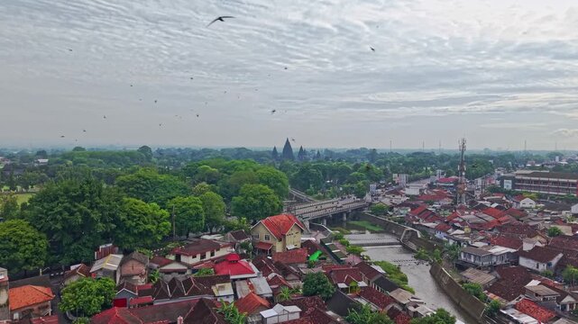Aerial view of the ancient Prambanan Temple contrasting with red-roofed buildings along a river, a captivating blend of history and urban life, Yogyakarta, Indonesia.
