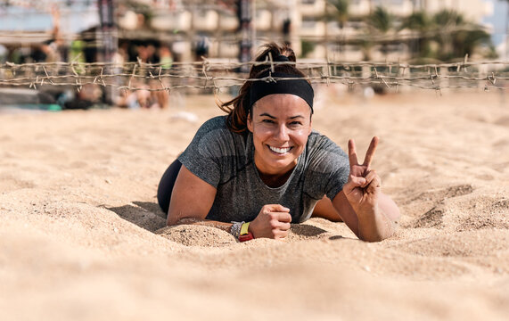 Happy female athlete crawling under barbed wire on the beach sand during an obstacle course, smiling and making the victory sign.