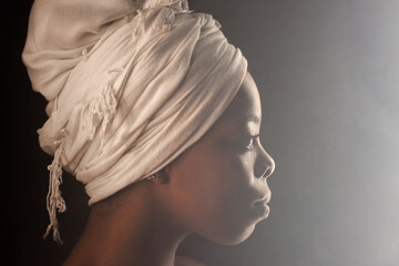 Profile portrait of attractive African American woman wearing white turban looking into the distance in fog on dark background