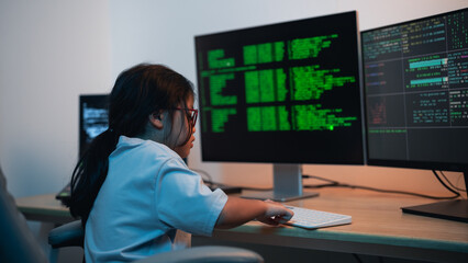 Young girl focused on coding and programming at computer desk with dual monitors displaying green...