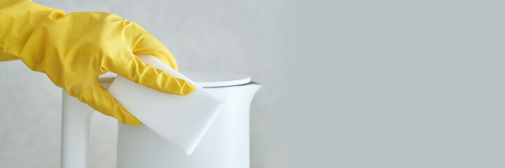 Gloved hand cleaning white kettle with sponge on light background.