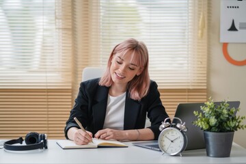 Woman with pink hair writing in notebook at desk with laptop and alarm clock in office setting