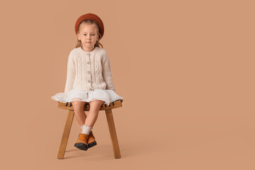 Stylish little girl in beret sitting on stool against beige background