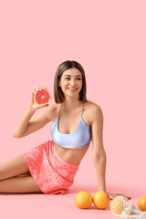 Young woman with grapefruits sitting on pink background