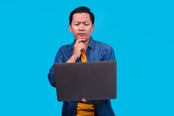 Pensive Young Asian Man Using Laptop Computer And Thinking Deeply Isolated On Blue Background